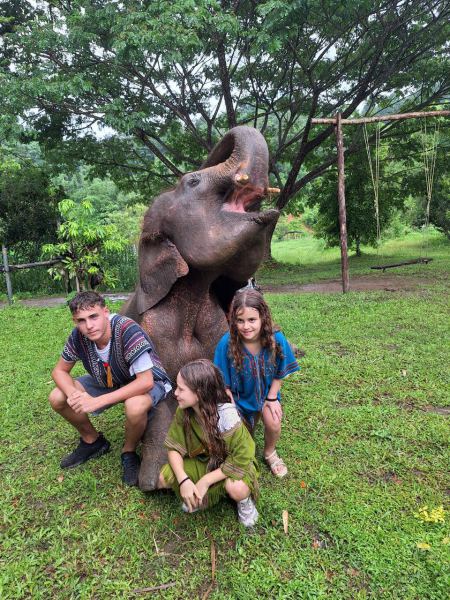 Three siblings kneeling beside a resting elephant with trunk raised - lifeonwheel.com
