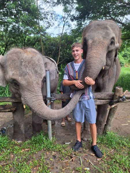 A young man standing between two elephants with trunks reaching to him - lifeonwheel.com